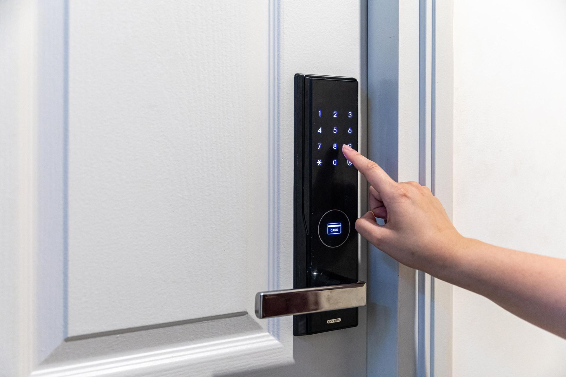 Hand entering code on a black digital door lock with a stainless steel handle on a white door.