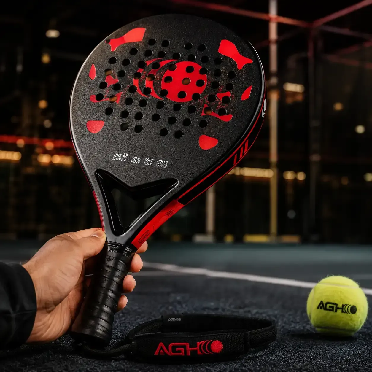 Hand holding a black and red AGH HO pickleball paddle beside a pickleball and paddle cover.