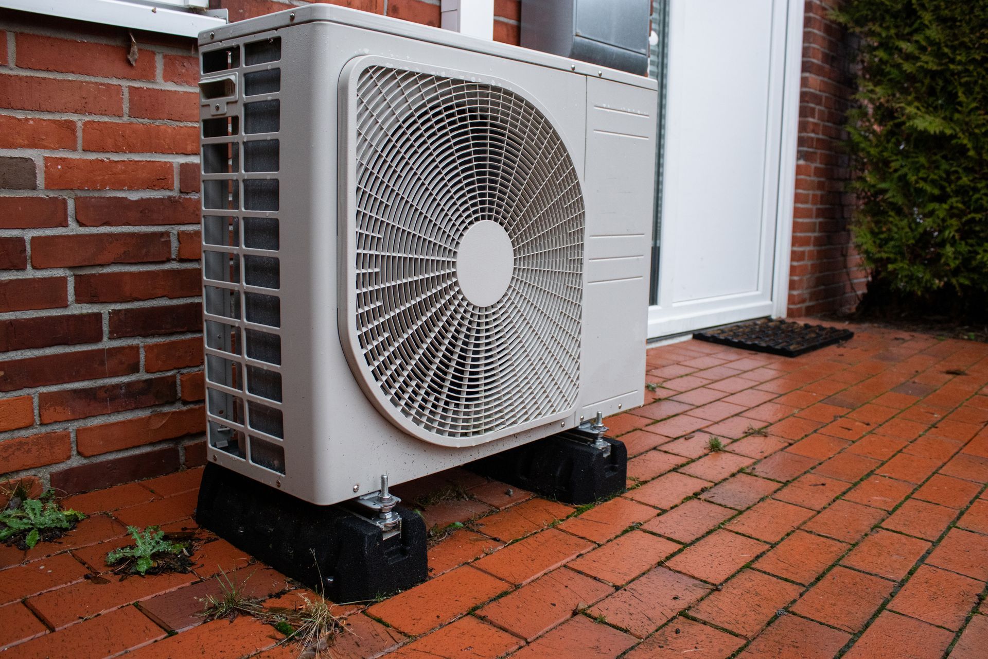 A beige outdoor heat pump unit mounted on black plastic risers on a red brick patio next to a brick wall.