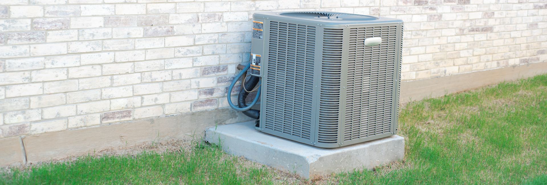 An outdoor air conditioning condenser unit sitting on a concrete pad against a light-colored brick exterior wall.