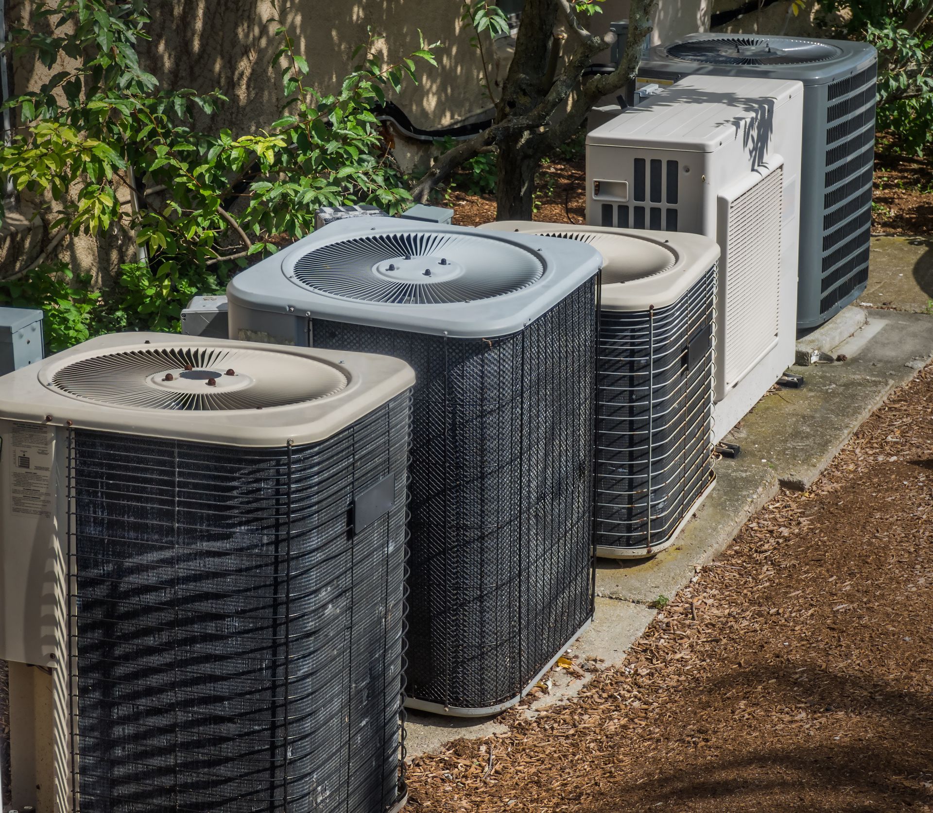 A row of four outdoor HVAC condenser units sitting on a concrete slab next to green foliage.
