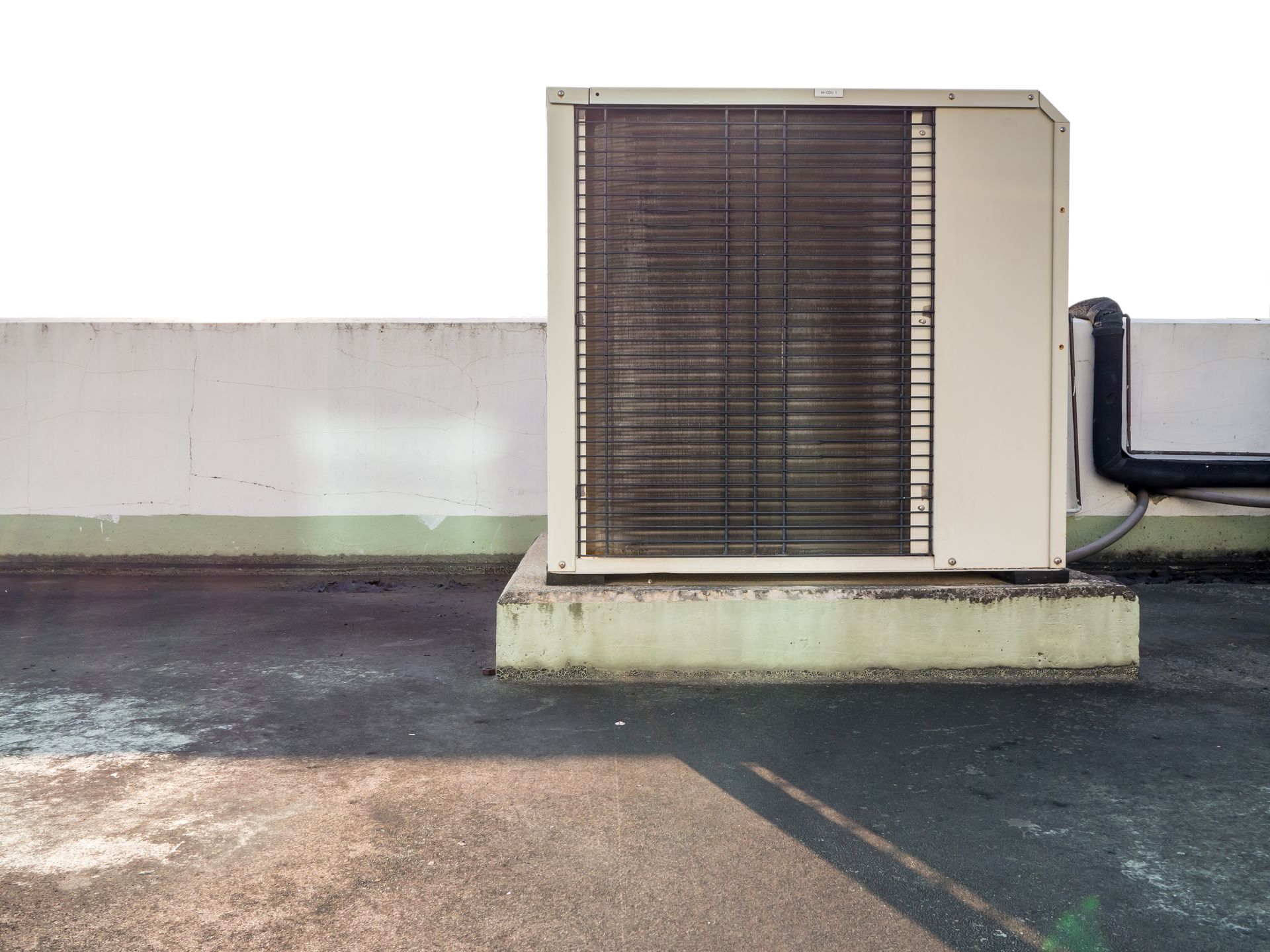 A beige air conditioning condenser unit sits on a raised concrete platform on a flat, weathered rooftop.