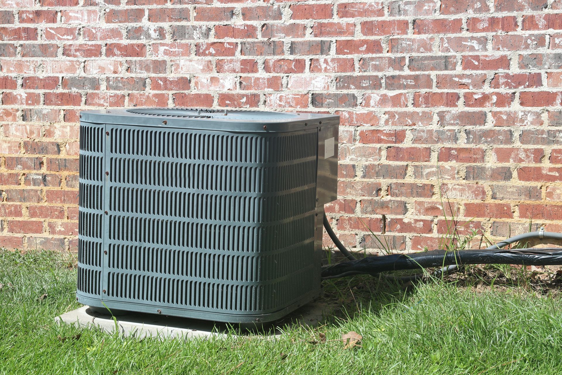 A gray outdoor air conditioning condenser unit sits on a concrete pad against a red brick wall in a grassy yard.