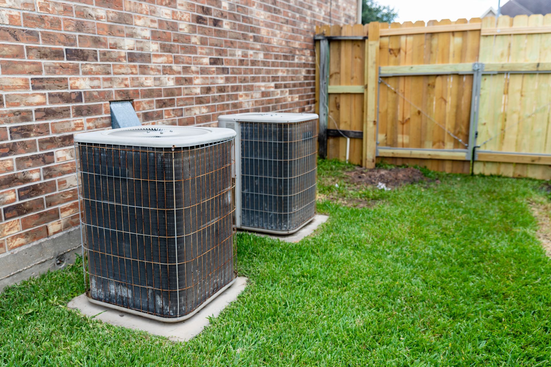 Two outdoor air conditioning units sit on concrete pads in a grassy backyard beside a brick wall and a wooden fence.