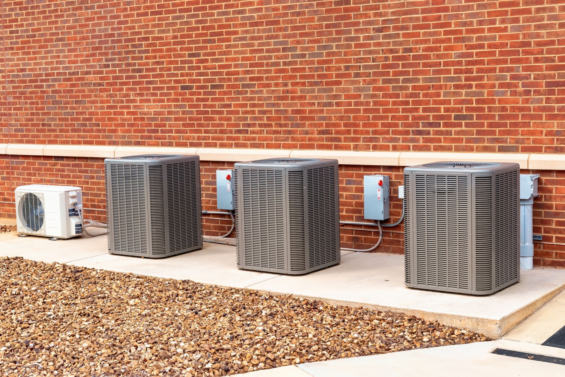 Three grey HVAC condenser units sit on a concrete pad against a red brick wall, with a smaller white unit on the left.