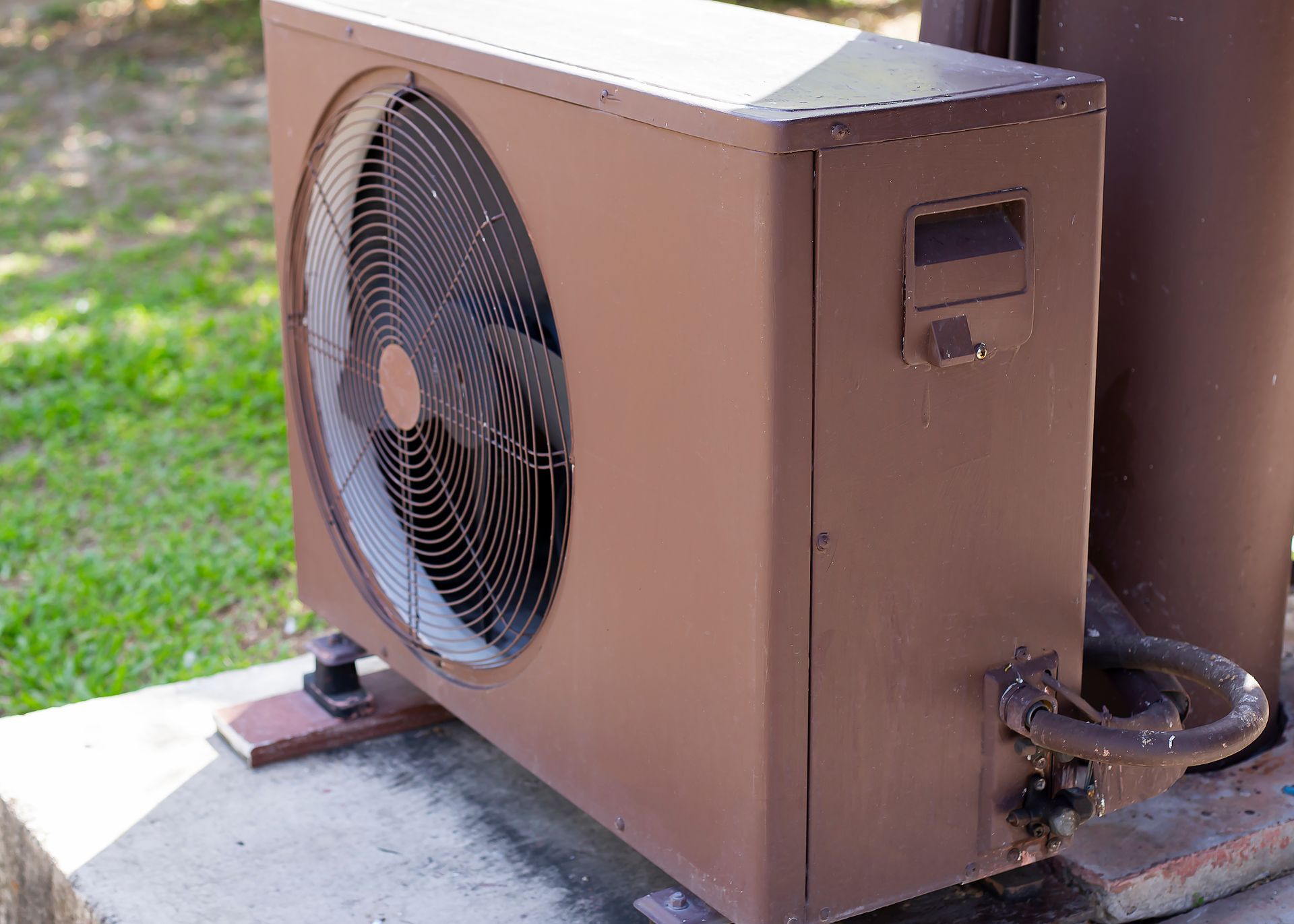 A brown outdoor air conditioning unit sits on a concrete slab in a grassy area.