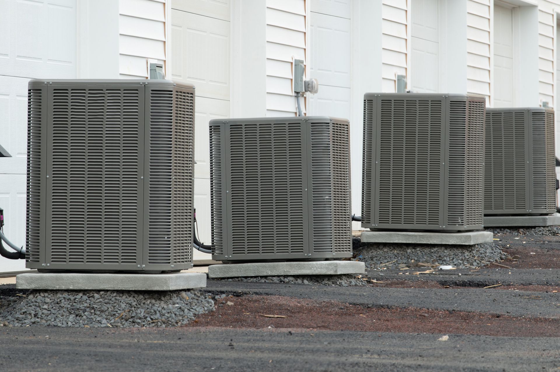 Four gray HVAC condenser units sit on concrete pads in a row along a white building wall.
