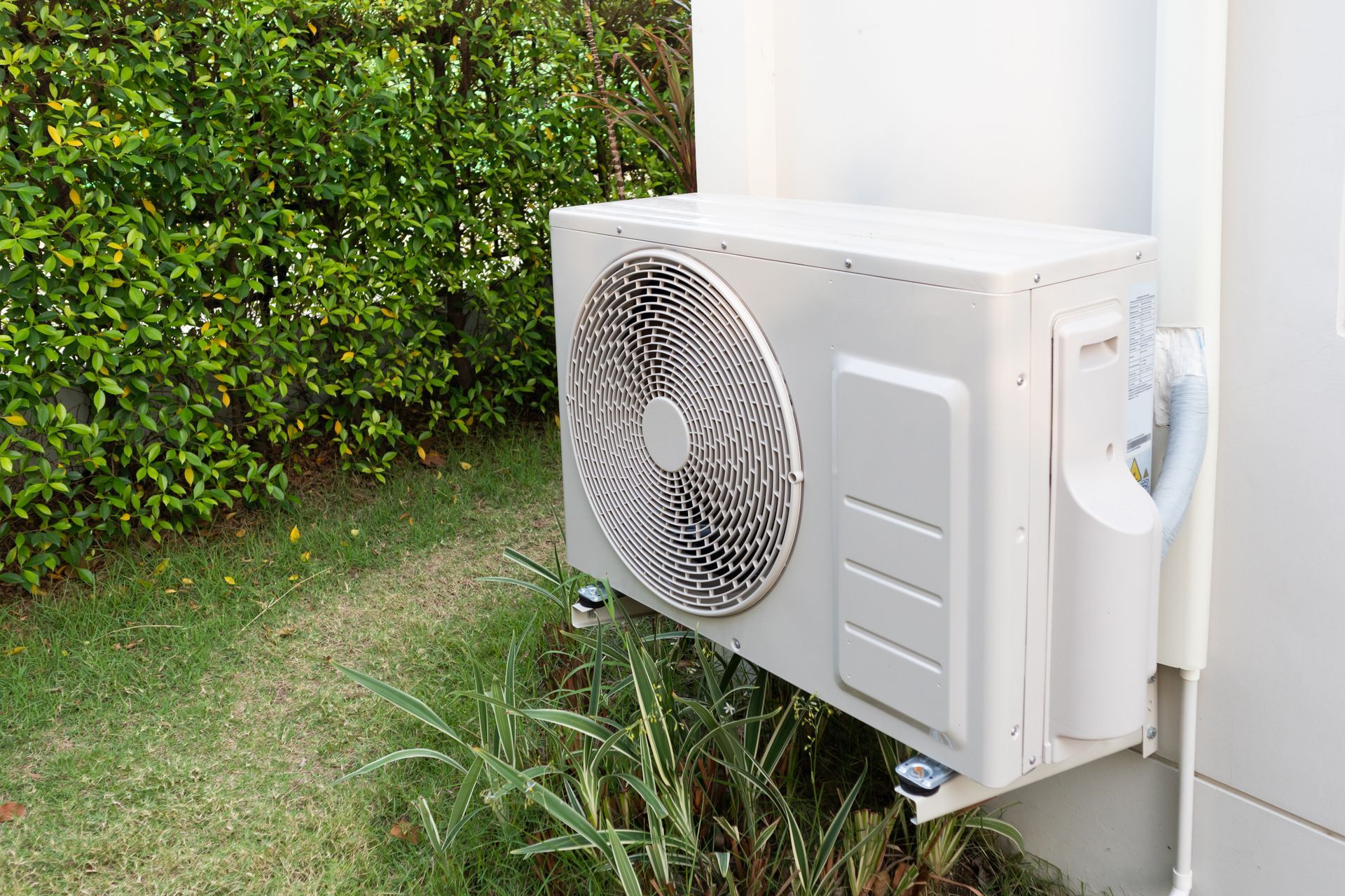 A light-colored outdoor air conditioning unit mounted on a white wall next to green foliage and grass.