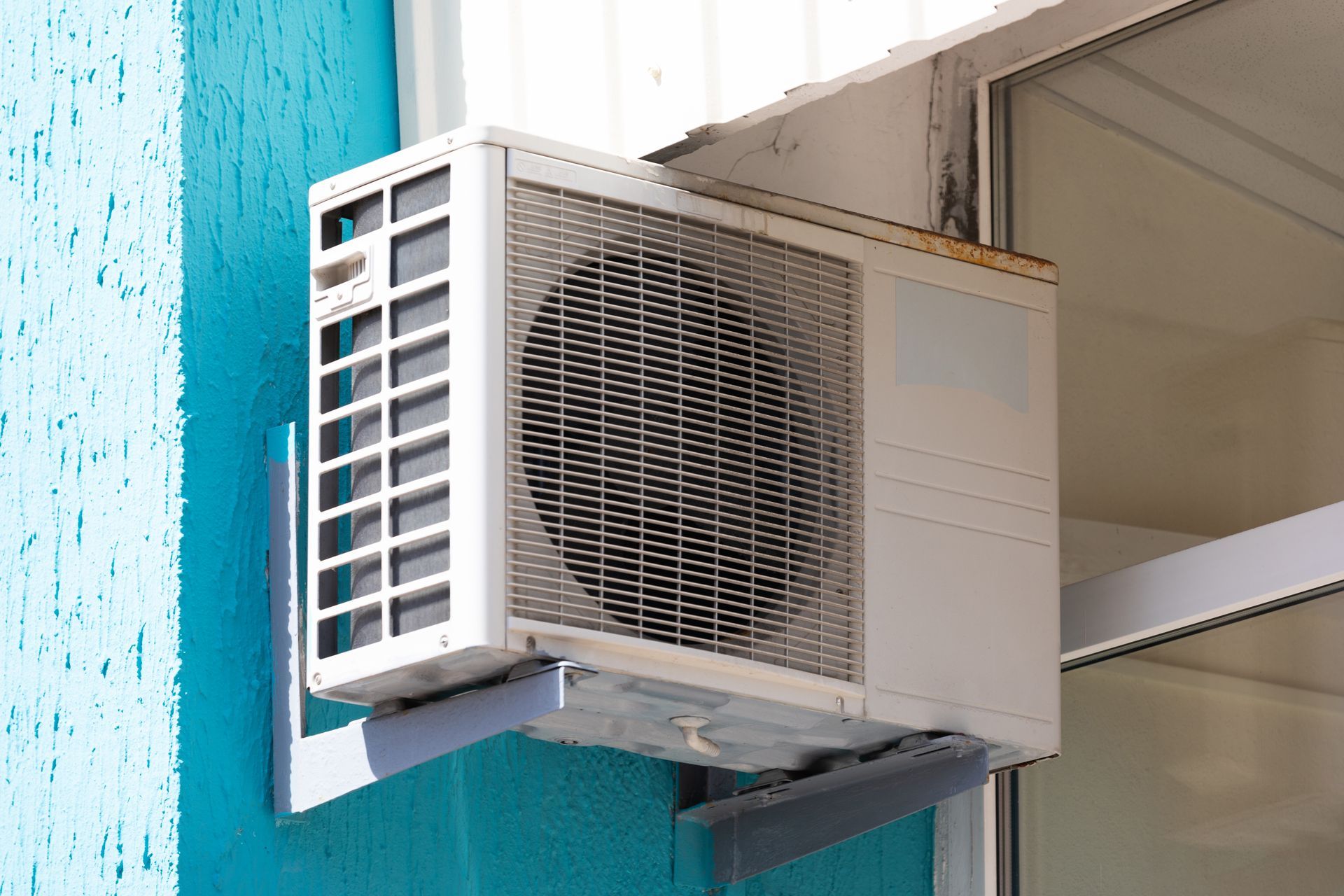 A white outdoor air conditioning unit mounted on a bright blue wall next to a window.