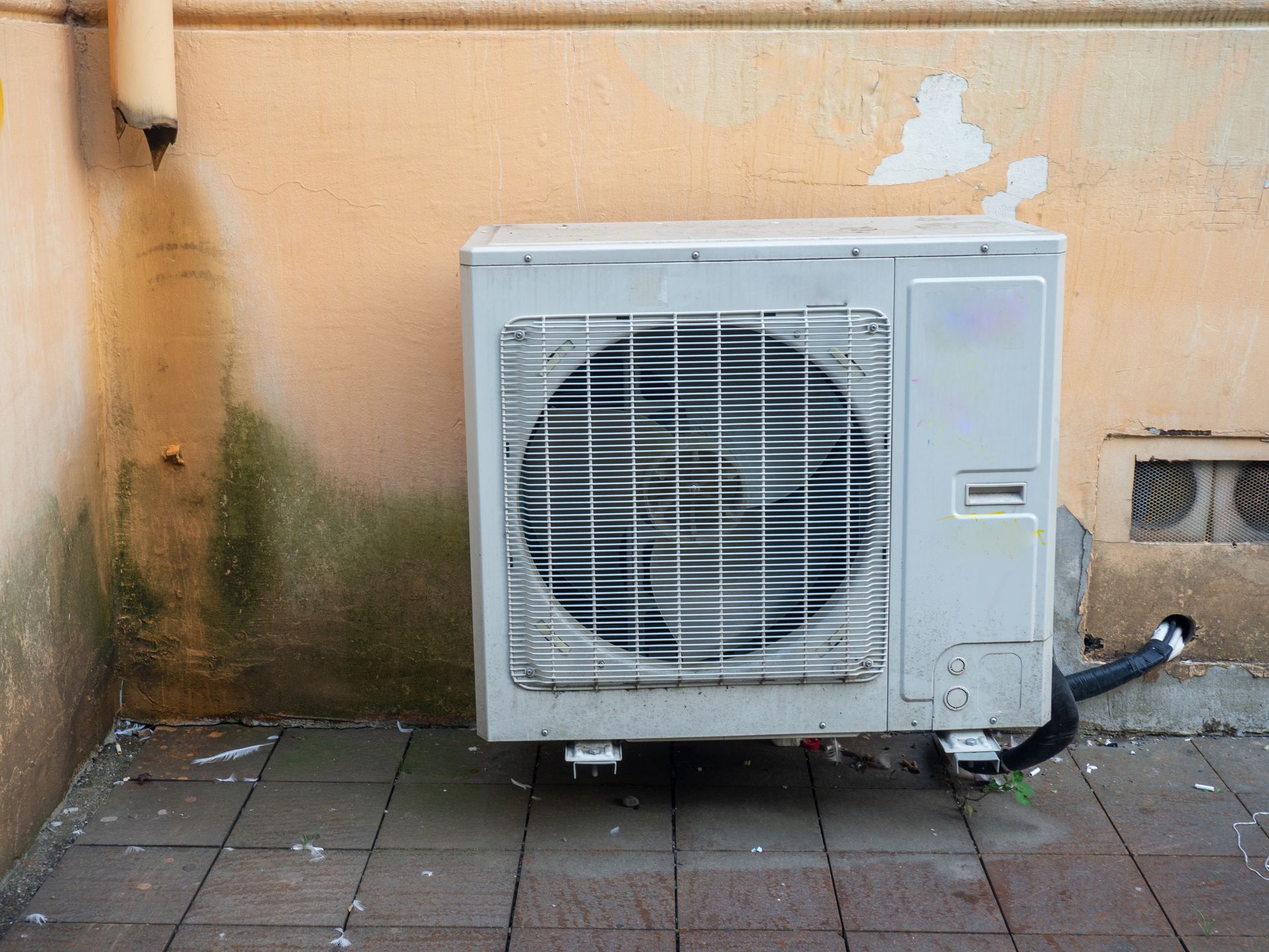 A white outdoor air conditioning unit sits on a tiled patio against a peach-colored wall with visible water staining.