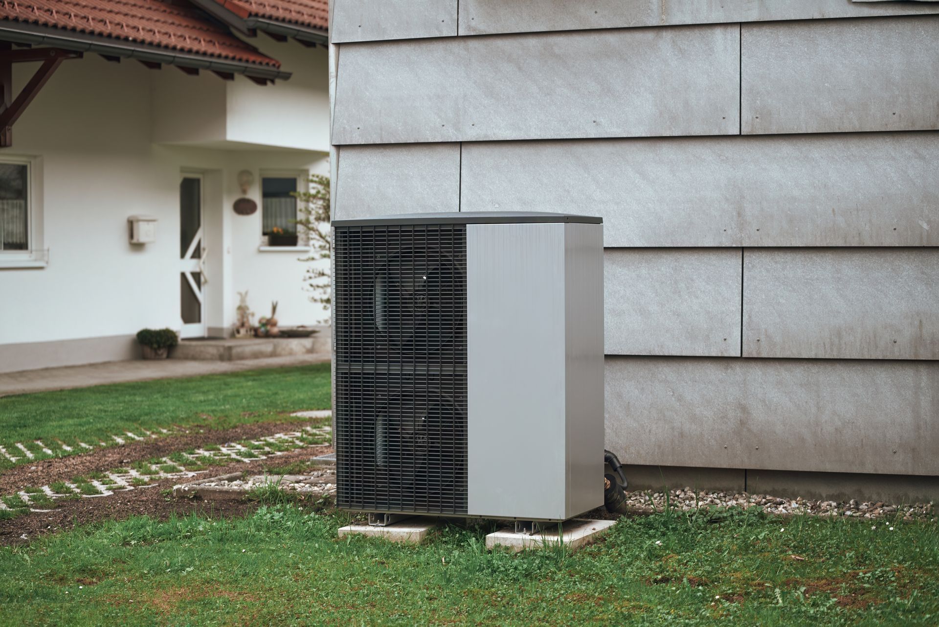 An outdoor heat pump unit sits on a concrete base next to a residential building wall with a house in the background.