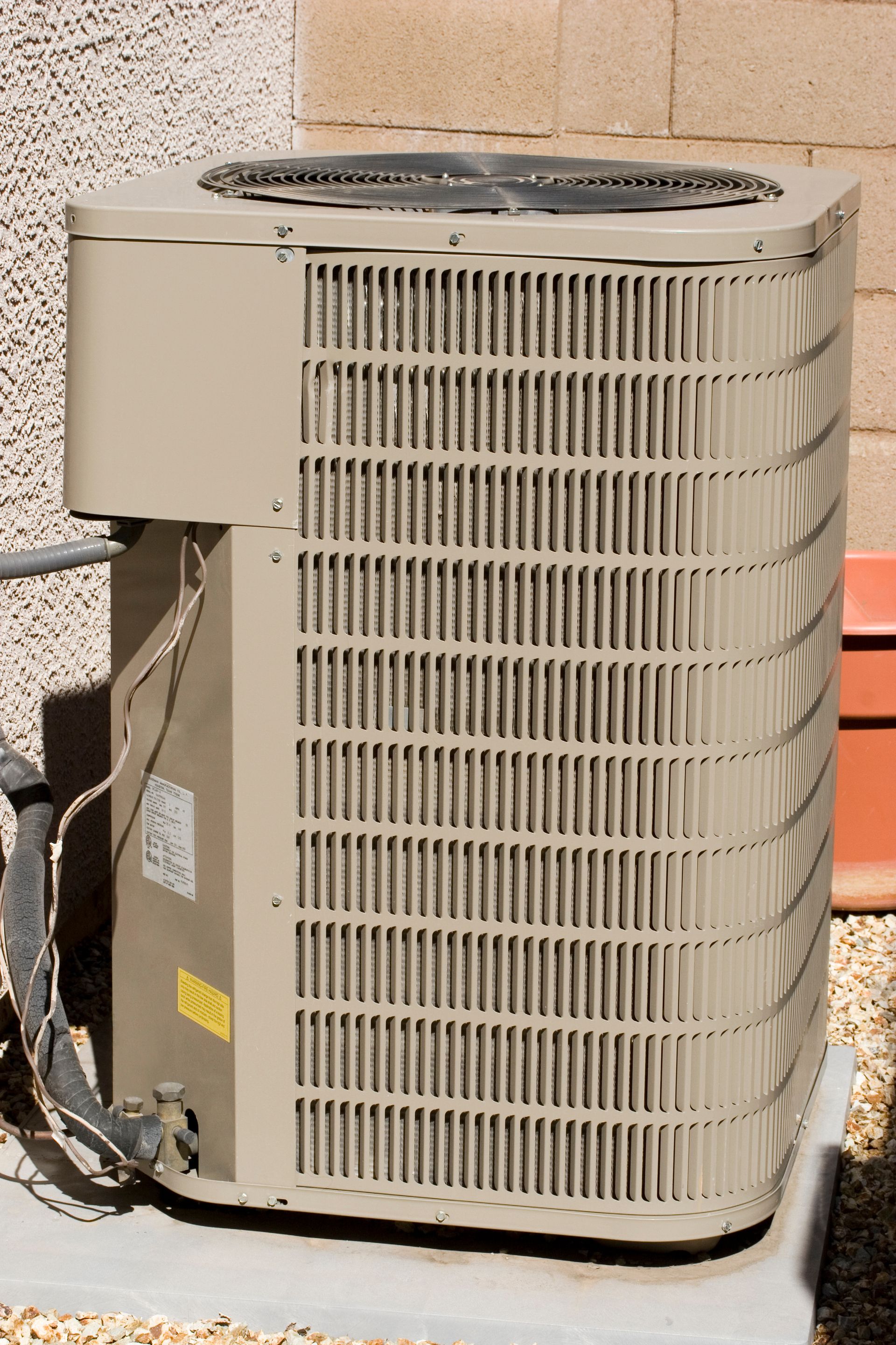 A beige, rectangular outdoor air conditioning unit sits on a concrete pad against a light-colored exterior wall.