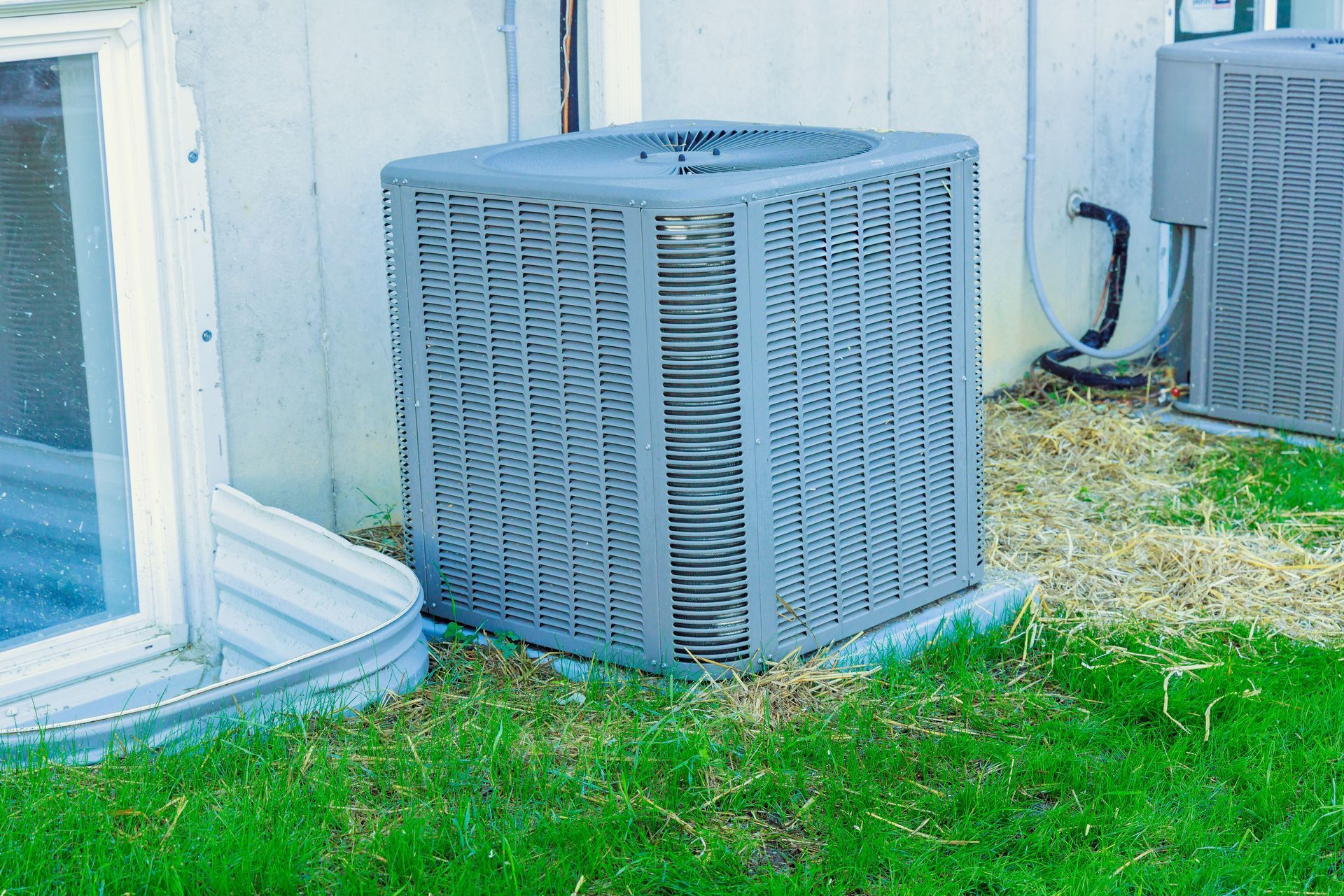 Two gray air conditioning units sitting on the grass outside the foundation of a building.