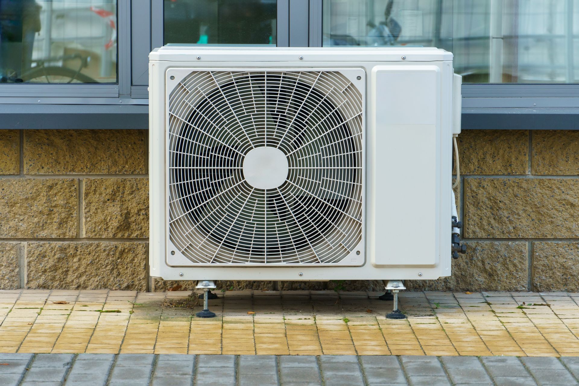 A white outdoor air conditioning unit sits on a paved patio against a brick wall and a window.