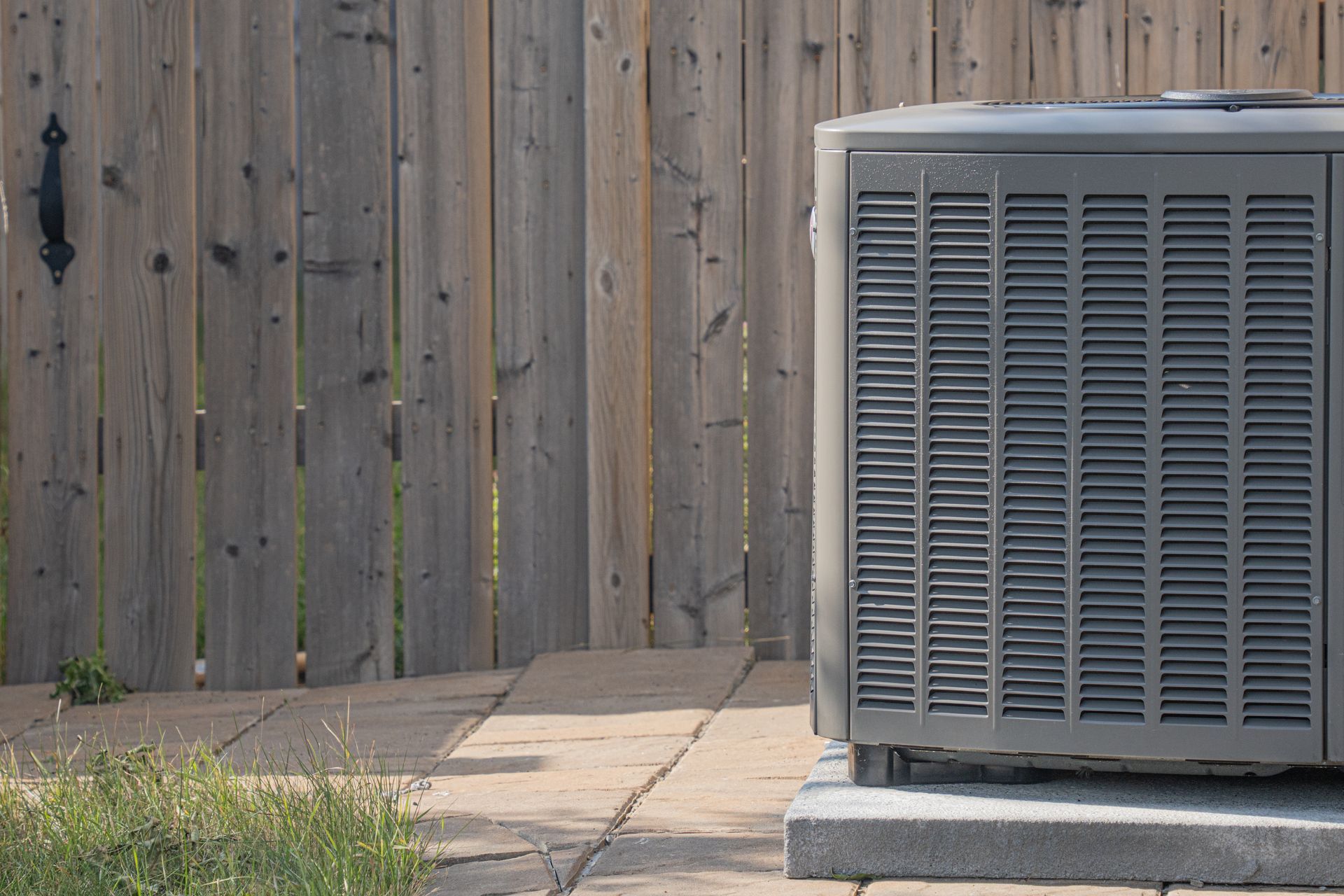 An outdoor air conditioning unit sits on a concrete pad next to a vertical wooden fence.