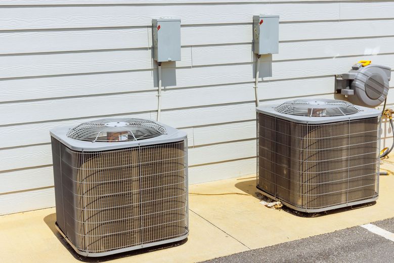 Two outdoor air conditioning units sit on a concrete pad against a house with light-colored horizontal siding.