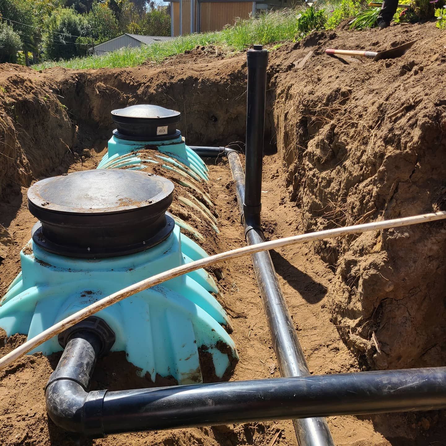 Two turquoise septic tanks in a trench, connected by black pipes, earth surrounding.