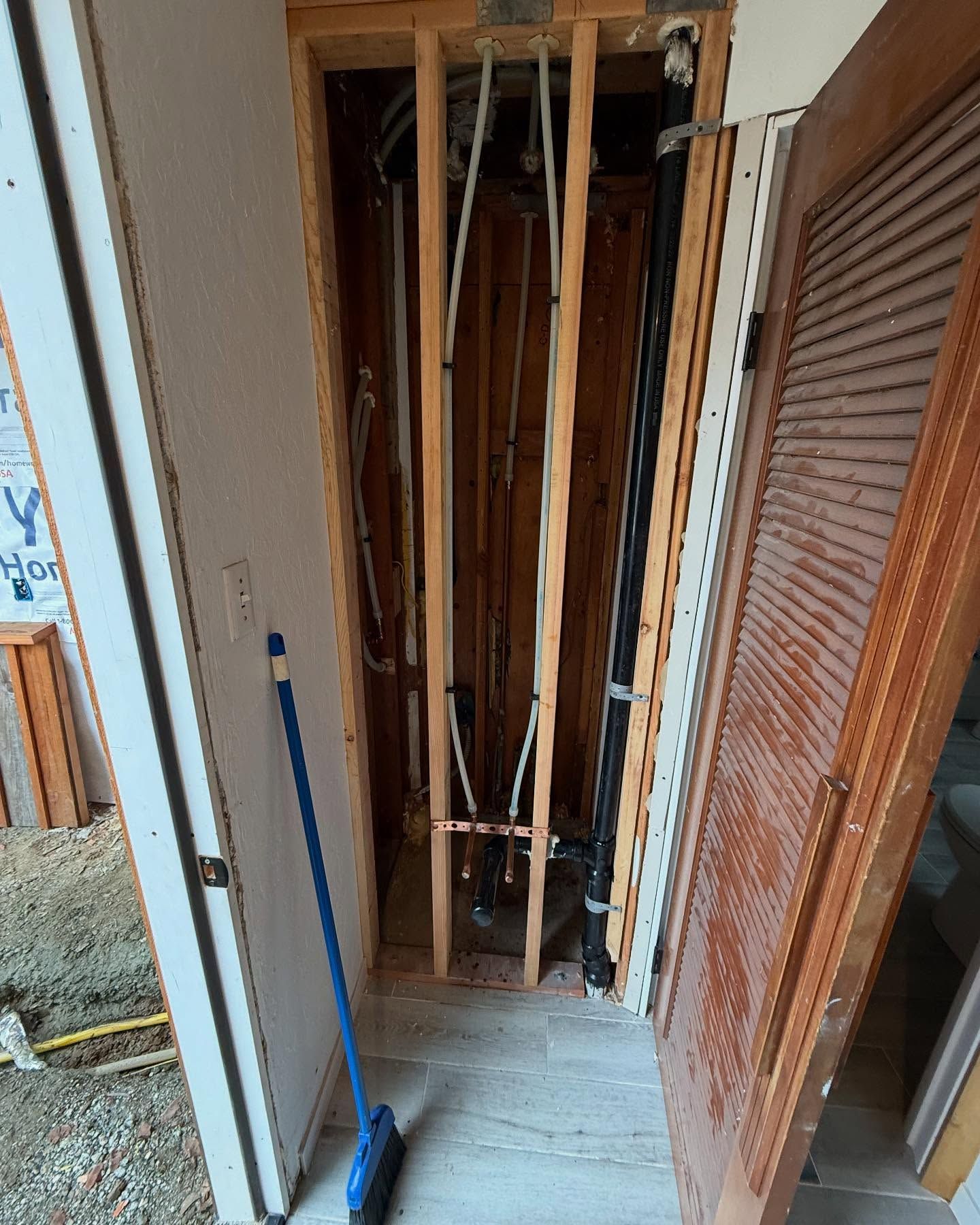 Framed doorway reveals plumbing and pipes in a wall. Broom leans against the wall.