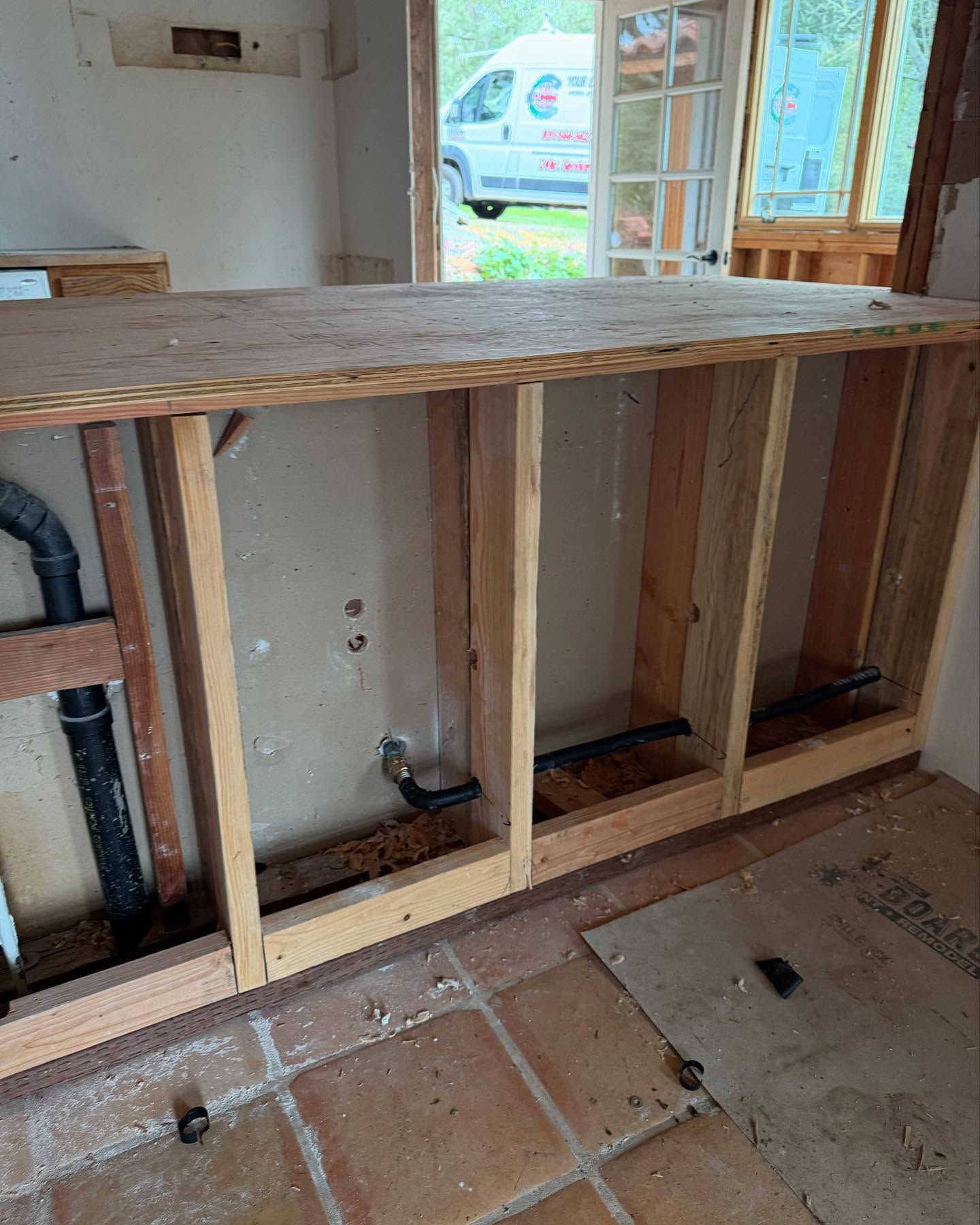 Interior view of a kitchen under renovation, showing exposed wooden frame and plumbing behind a countertop.