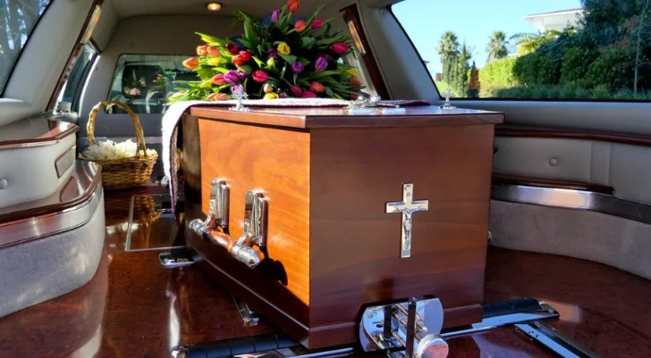 Wooden casket with white cross and flowers inside a hearse trunk