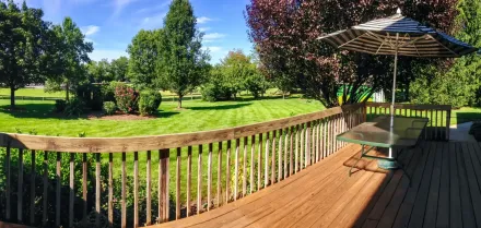 Sunlit wooden deck overlooking a green lawn and trees, with a patio umbrella and chair.