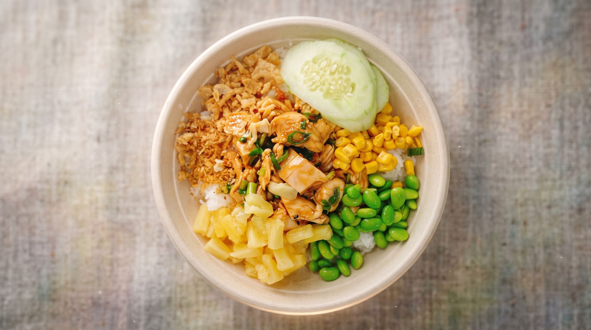 Bowl of rice with sliced chicken, cucumber, corn, peas, and pineapple on a wooden table