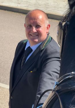 Smiling bald man in a dark suit with a striped tie standing outdoors in sunlight