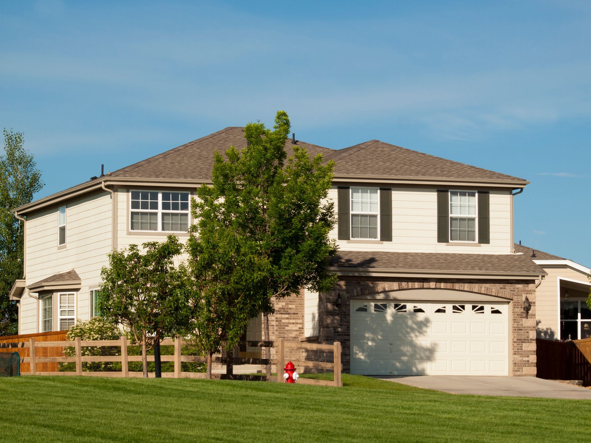 Suburban two-story house with beige exterior, brown roof, garage, and a small tree in front