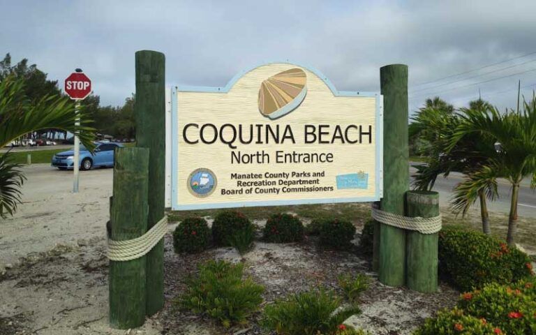 Coquina Beach North Entrance sign with rope posts, palm plants, and a stop sign in the background