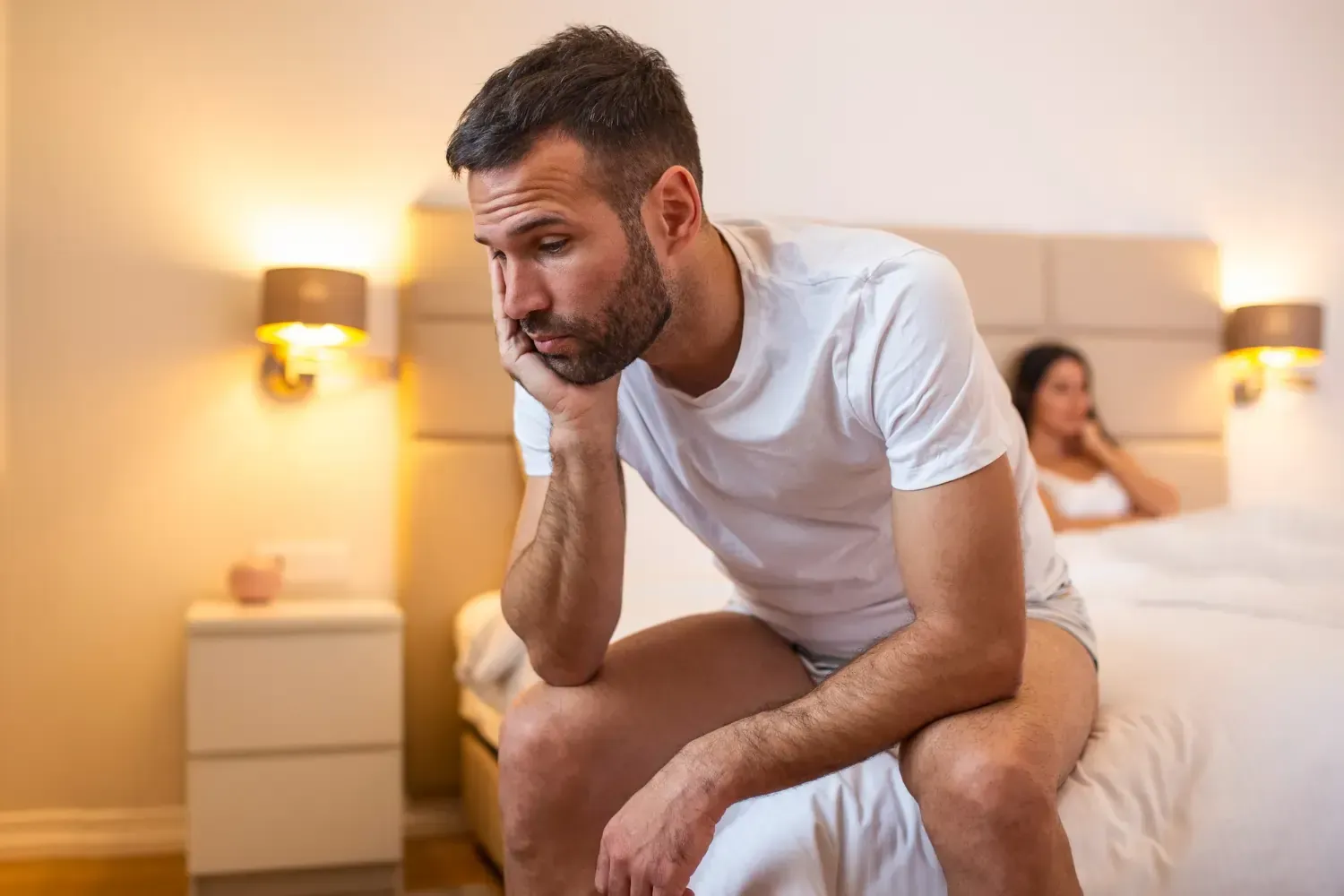 A man is sitting on a bed with a woman in the background.