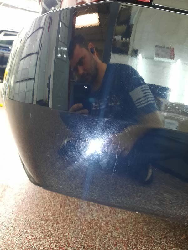 Person taking a photo reflected in a black car bumper covered in swirl marks, indoors.