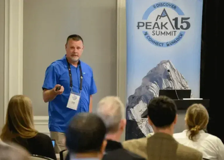 Speaker presenting at PEAK 15 Summit beside mountain banner, facing an audience in a conference room