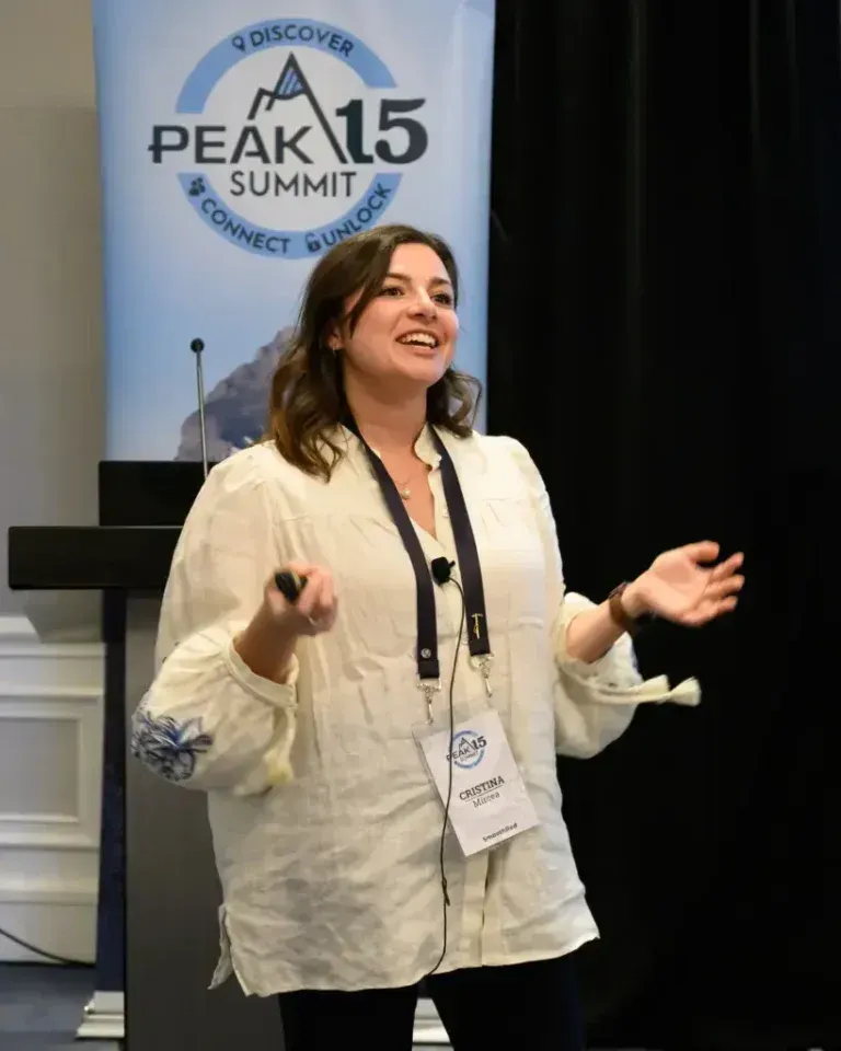 Woman speaking onstage in a white blouse, gesturing before a Peak 15 Summit banner.