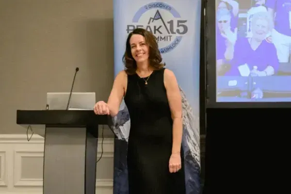 Woman smiling beside a podium at a conference stage with a BEAK15 banner and projected screen behind her