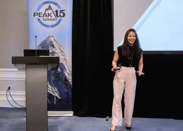 Woman speaking onstage beside a podium and “PEAK 15 SUMMIT” banner, smiling with a remote in hand