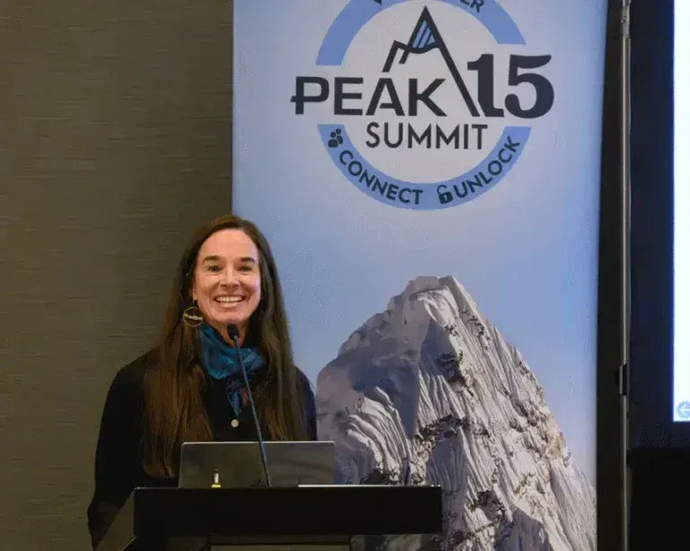 Speaker at a podium beside a PEAK Summit 15 banner, smiling in a conference room.