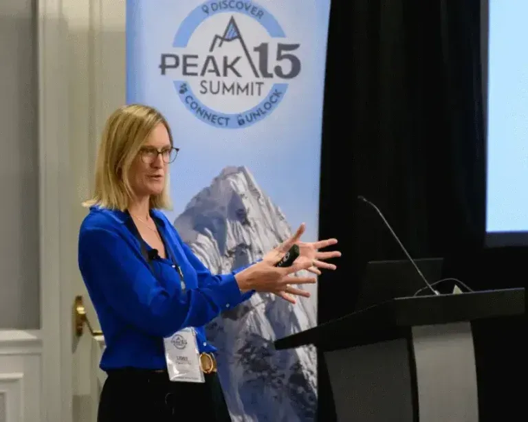 Speaker in blue jacket presenting at Peak15 Summit podium beside mountain banner