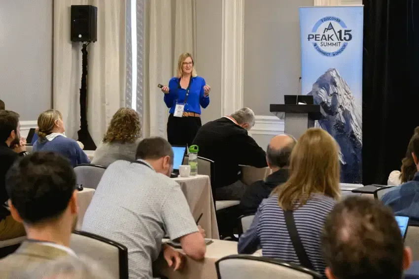 Speaker presenting to seated audience in a conference room, with a PEAK15 mountain banner behind her.