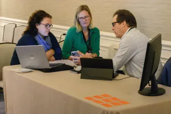Three coworkers discussing at a table with laptops and a monitor in a meeting room.