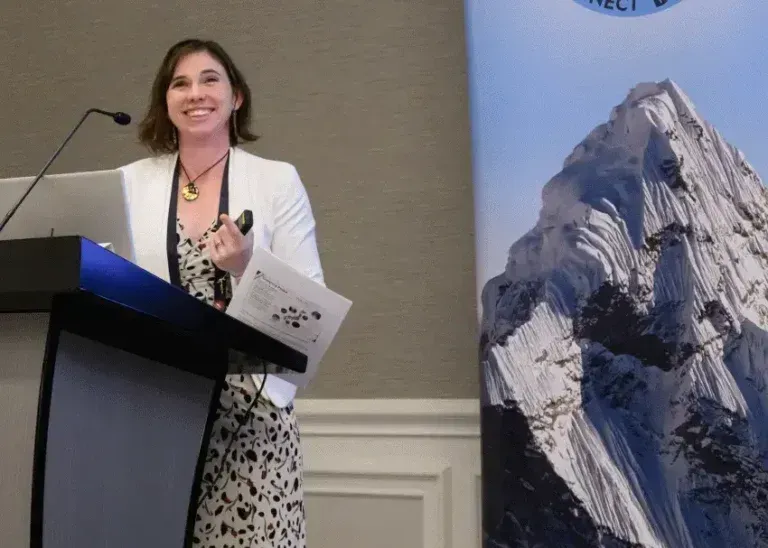 Woman speaking at a podium beside a mountain poster, holding papers and smiling.