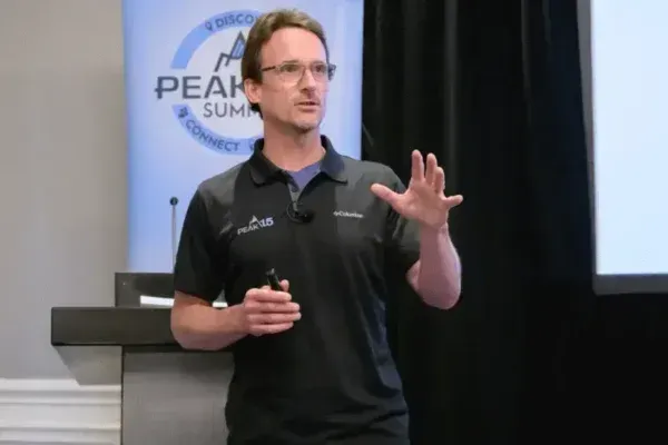 Presenter in black polo speaking at a conference podium, gesturing in front of a Peak... summit backdrop