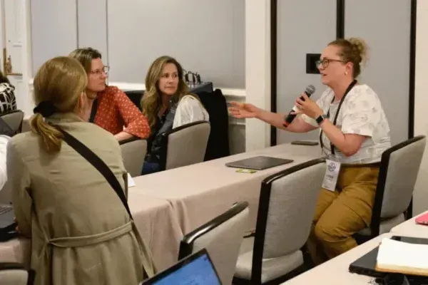 People seated at a conference table listening to a woman speaking into a microphone.