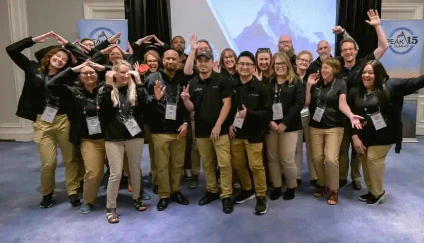 Group of people in black shirts and khaki pants posing with raised arms at a conference booth