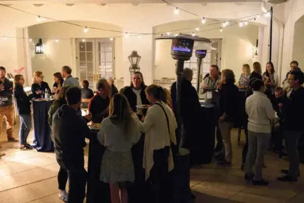 People gathered at an indoor evening reception under warm lights, mingling around a central bar.