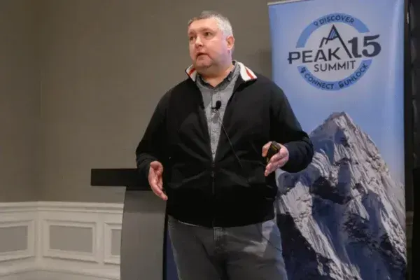 Man speaking on stage beside a Peak 15 Summit banner with a mountain image.