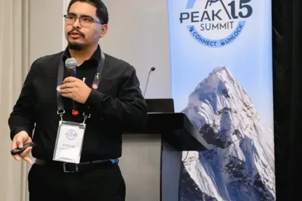 Speaker holding a microphone at Peak 15 Summit beside a mountain banner