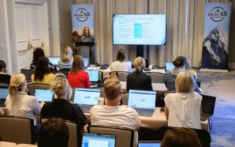 Conference audience seated at laptops, watching a presenter speak beside a large screen in a hotel meeting room.