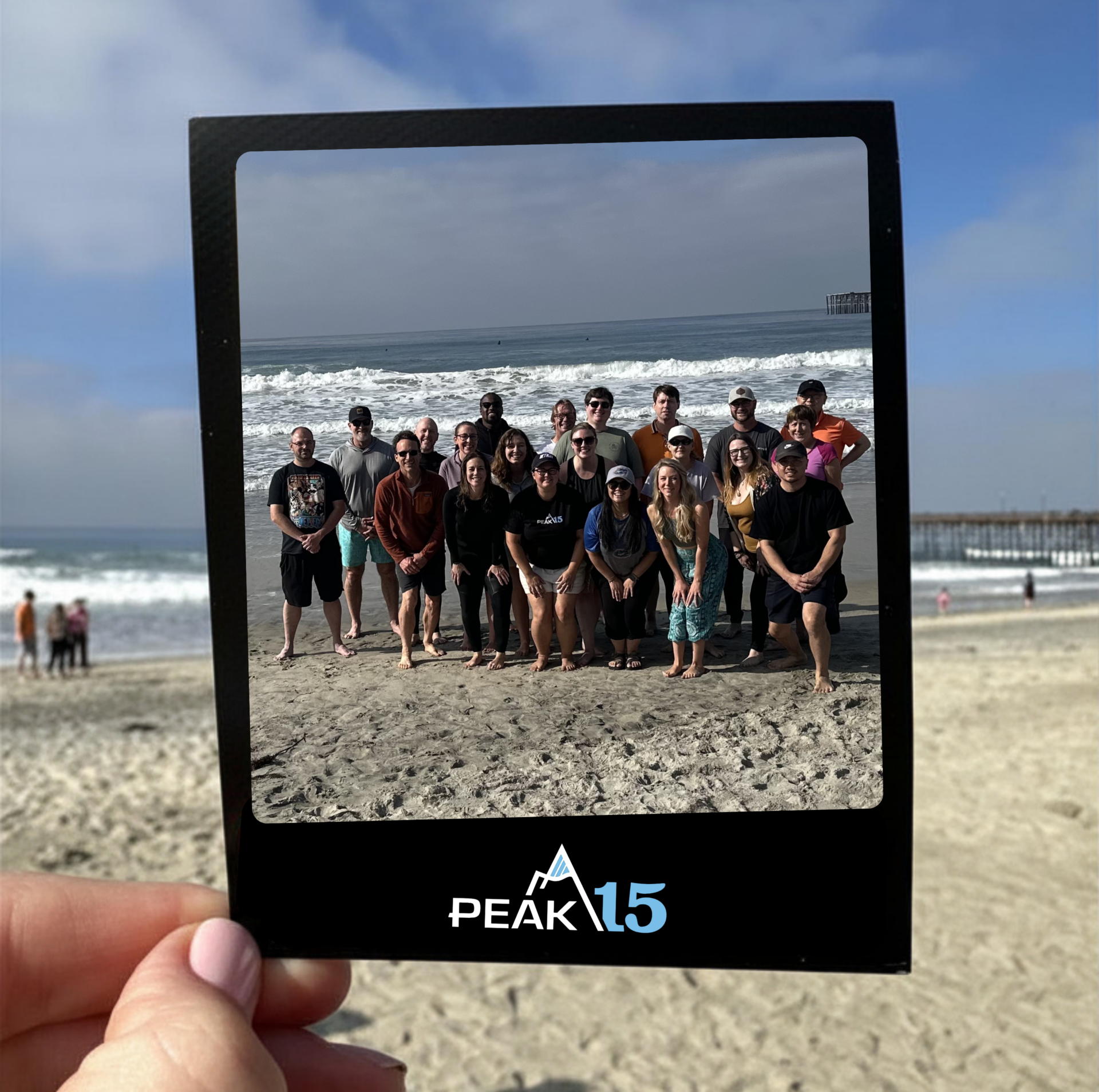 Hand holding a photo frame showing a group posing on a beach with waves behind them