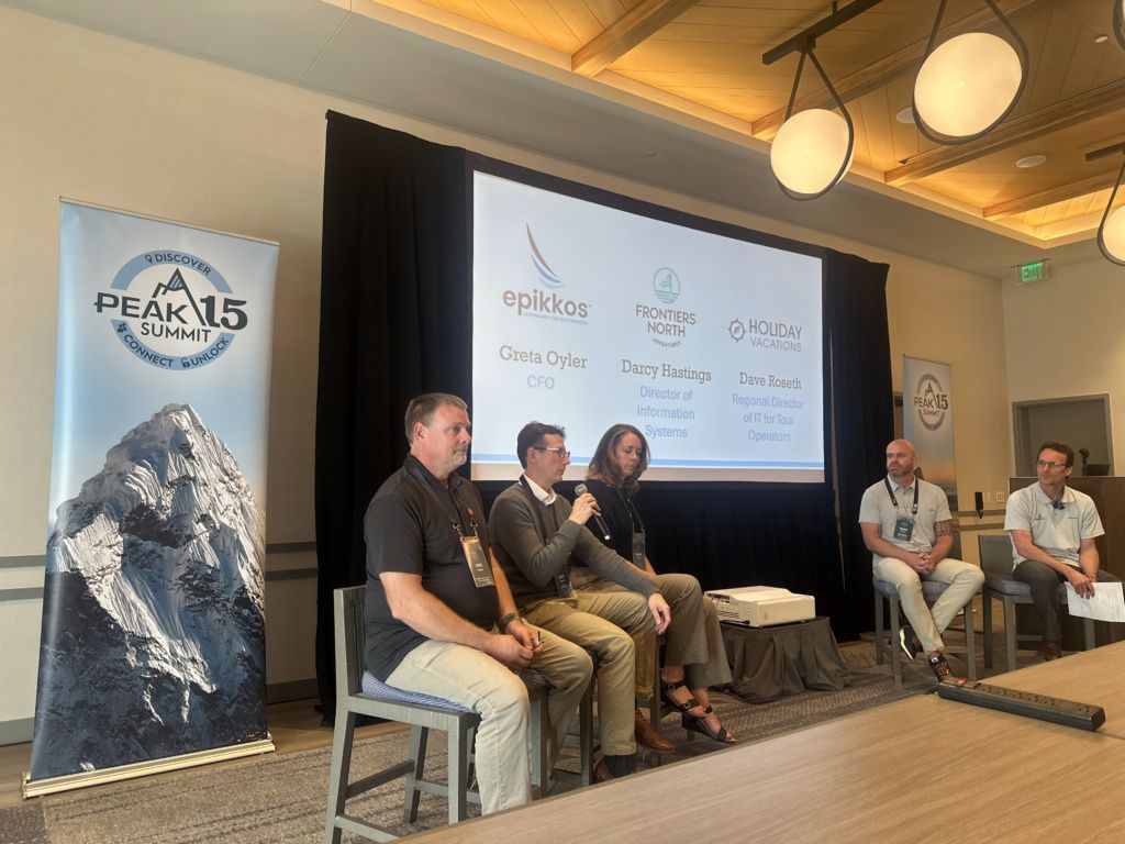 Panel discussion on stage with five seated speakers, sponsor banner, and projected presentation screen
