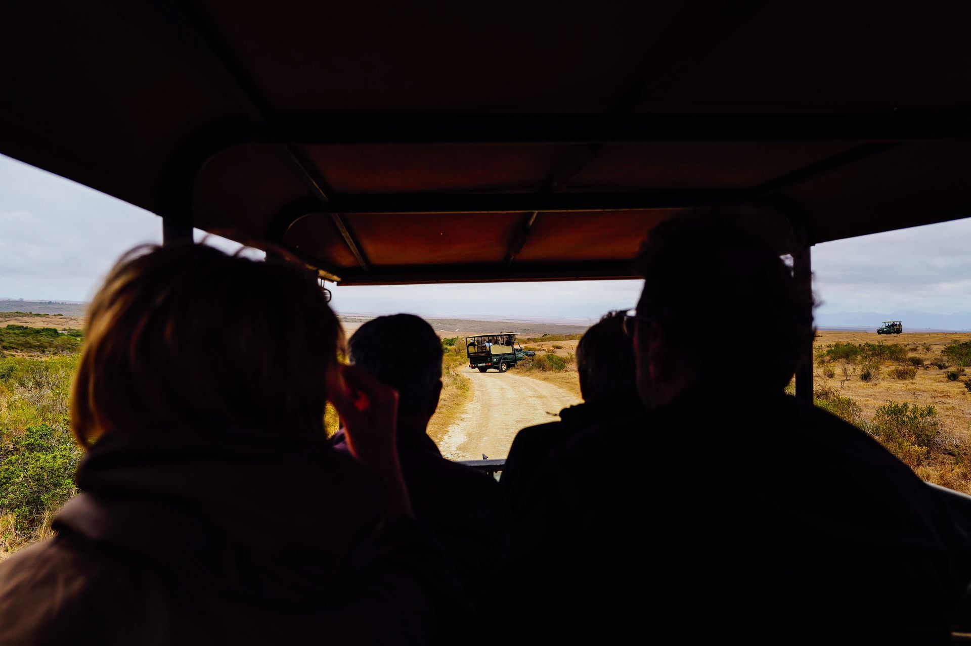 Passengers ride in an open safari vehicle on a dirt road, following another vehicle through a dry landscape.